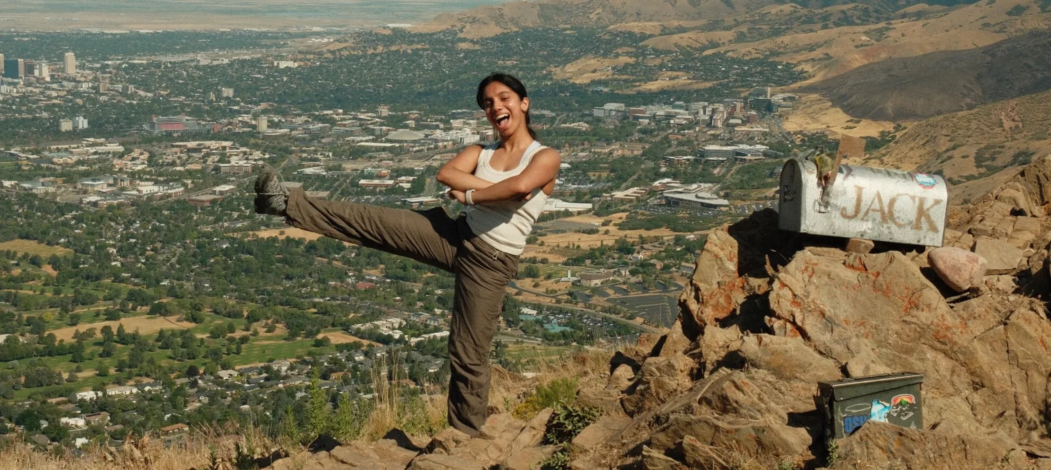 Shradha mid-kick on a rocky summit beside a mailbox painted JACK, with the city spread out in the valley below.